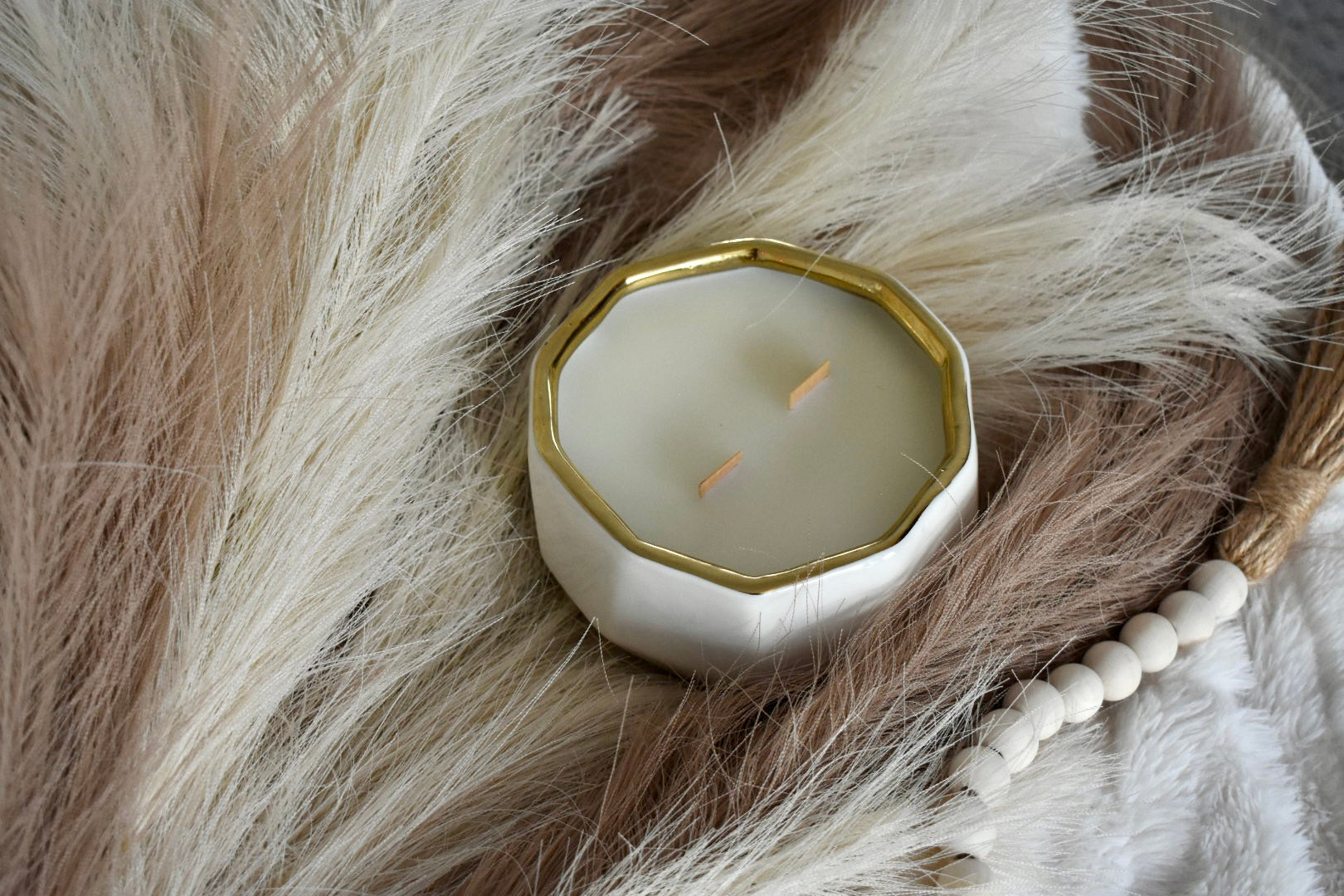 White candle jar with gold rim and gold interior. Coconut wax and wood wick. Displayed on a lush feather